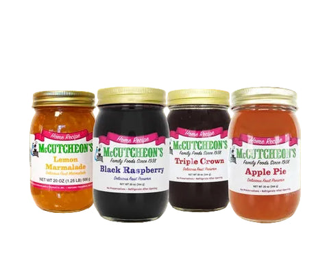 Four jars of McCutcheon's Family Foods gourmet fruit preserves arranged on a wooden surface with a blurred outdoor garden background. The jars include Lemon Marmalade, Black Raspberry, Triple Crown, and Apple Pie.