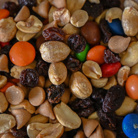 A close-up pile of mixed nuts and dried fruit, including tan peanuts, m&ms, raisins and peanut butter nibs