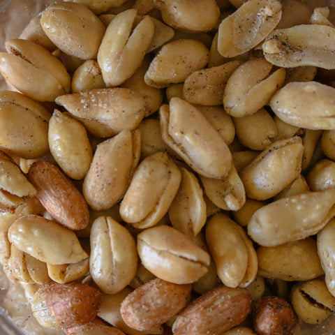 A close-up view of roasted peanuts in a clear container, showing their golden-brown, lightly toasted skins with some darker roasted pieces mixed in.
